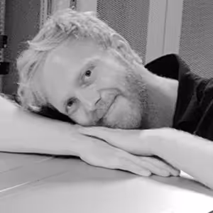 Black and white photo of a man with blond hair resting his head on his crossed arms on a table, smiling gently.