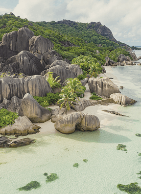 Tropical beach with large granite boulders, palm trees, and clear shallow water near a green hillside.