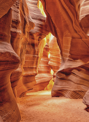Sunlit narrow sandstone canyon with smooth, layered walls and sandy floor.