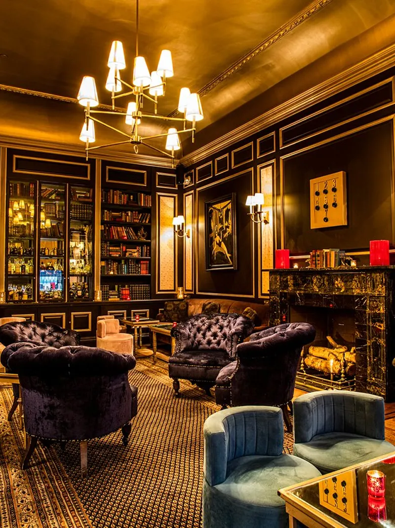 Luxurious lounge with velvet armchairs, ornate fireplace, bookshelf, and warm lighting.