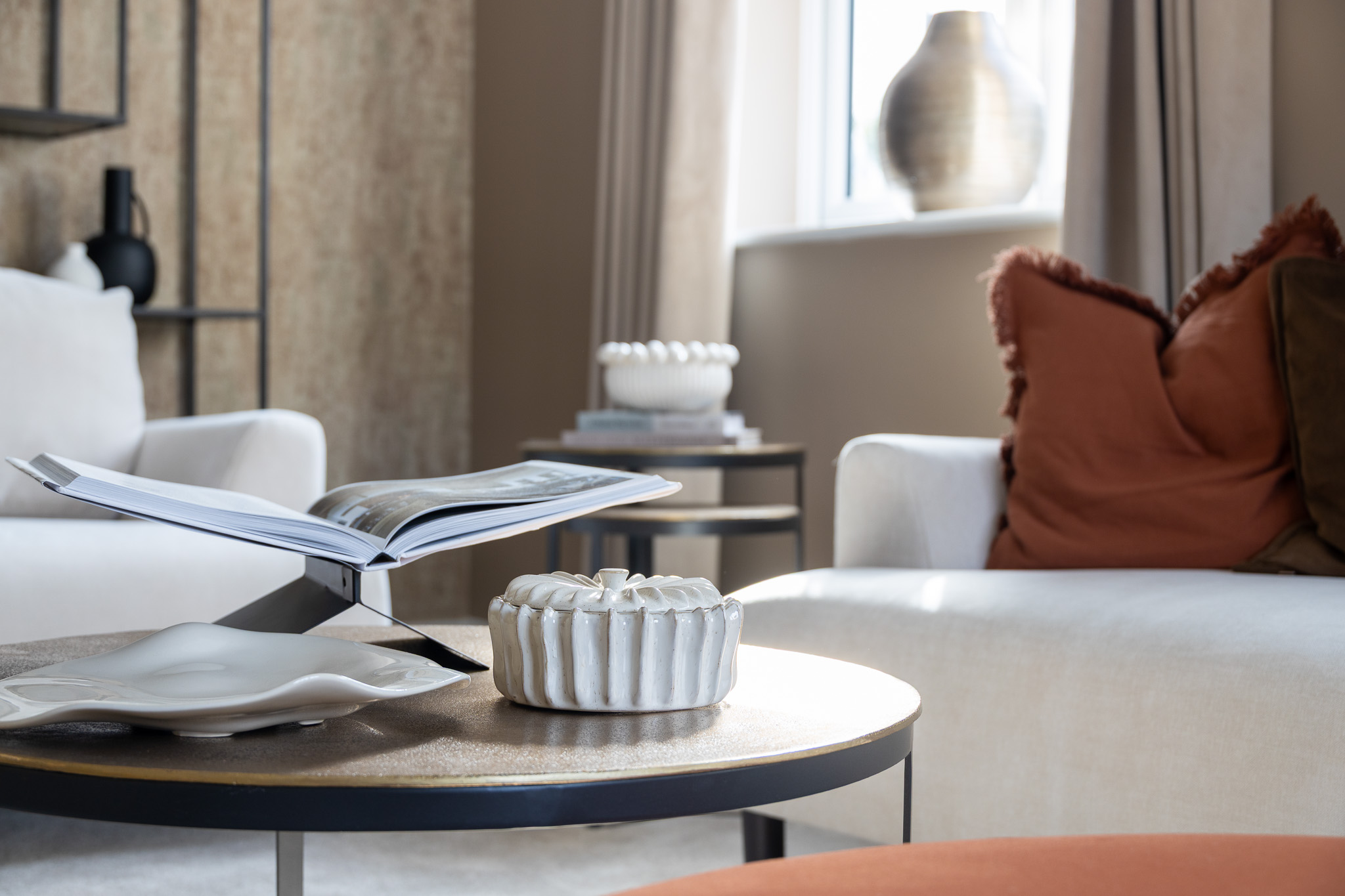 A stylish and cosy interior design shot of a living room. In the foreground, a round coffee table is styled with decorative objects, including an open coffee table book on a stand, a decorative white ceramic dish, and a small ribbed bowl with a lid. Two modern sofas with textured scatter cushions in terracotta and brown are visible in the background, near a window with sheer curtains that provide soft natural light. The room features a contemporary style with a warm colour palette.