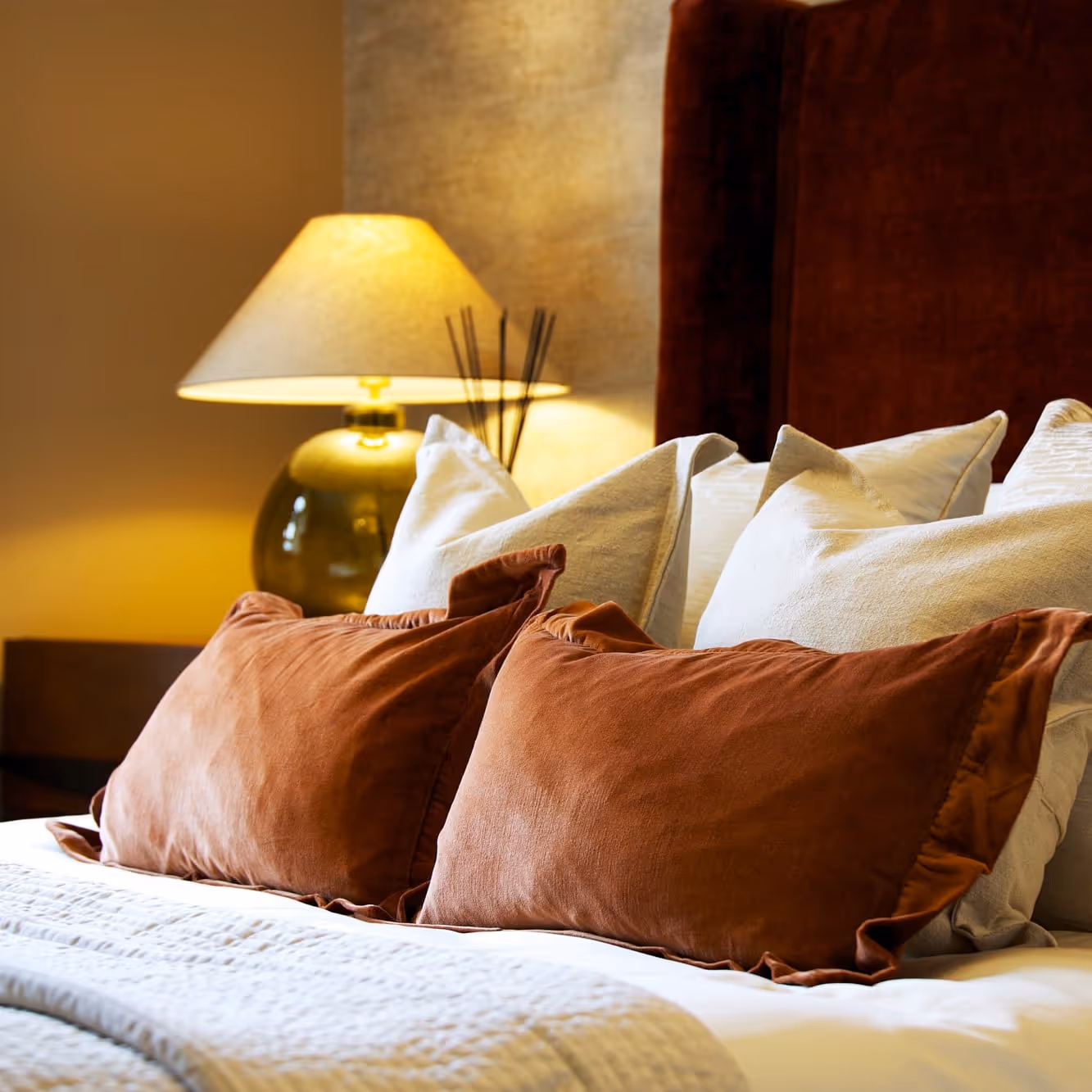 A close-up shot of a bedroom interior showcasing a luxurious bedding arrangement. A tall, plush headboard in a rich, warm tone is visible behind a stack of decorative scatter cushions in white and terracotta. On a bedside table, a modern table lamp casts a soft light, contributing to the cosy and comfortable atmosphere of the room. The design features a warm colour palette.