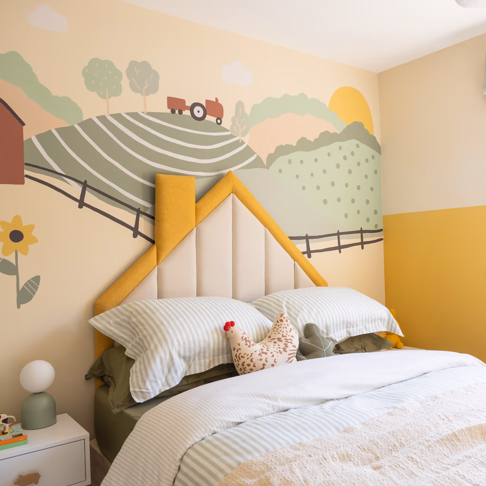A cheerful children's bedroom interior with a hand-painted farm-themed mural on the wall. The bed features a unique, house-shaped upholstered headboard and is dressed with white and striped bedding. A chicken soft toy sits on the bed. A window with a striped Roman blind lets in natural light, and a small bedside table with a lamp is visible. The room has a bright and cosy nursery design feel.