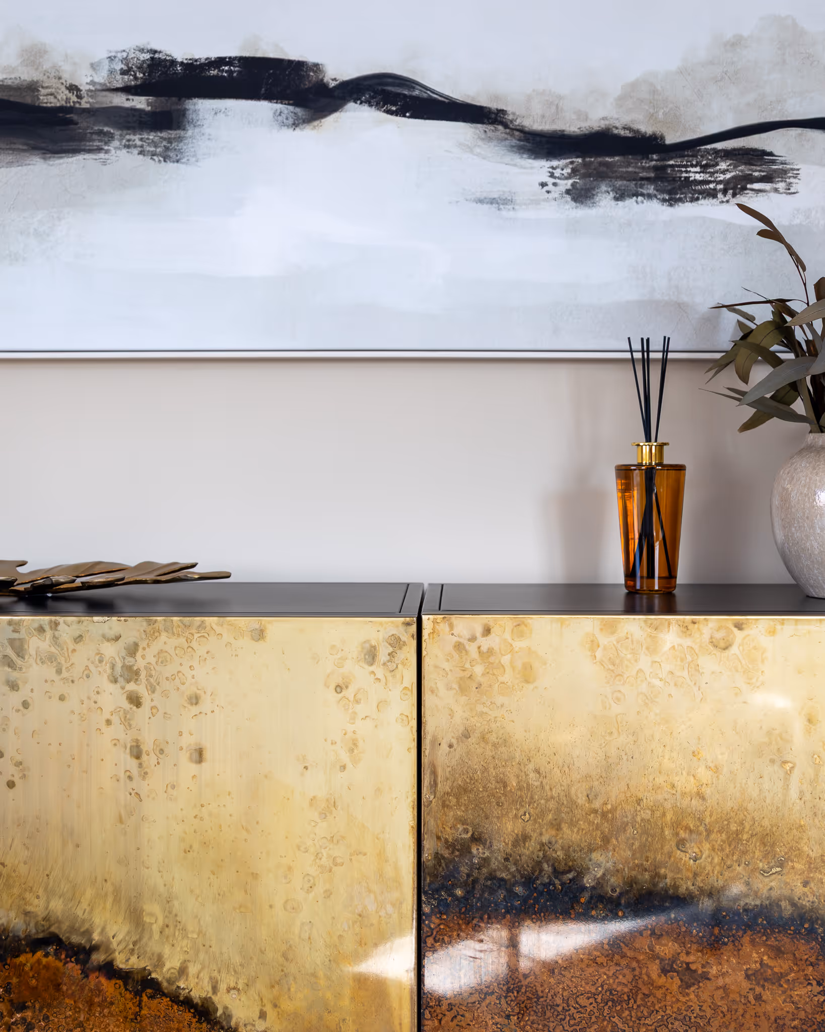 A close-up shot of a contemporary interior detail. The focus is a stylish sideboard with a unique, patinated brass finish on its doors. The surface is styled with a brown glass reed diffuser and a decorative vase holding dried leaves. Above the sideboard hangs a large piece of black and white abstract wall art, adding to the modern home decor.