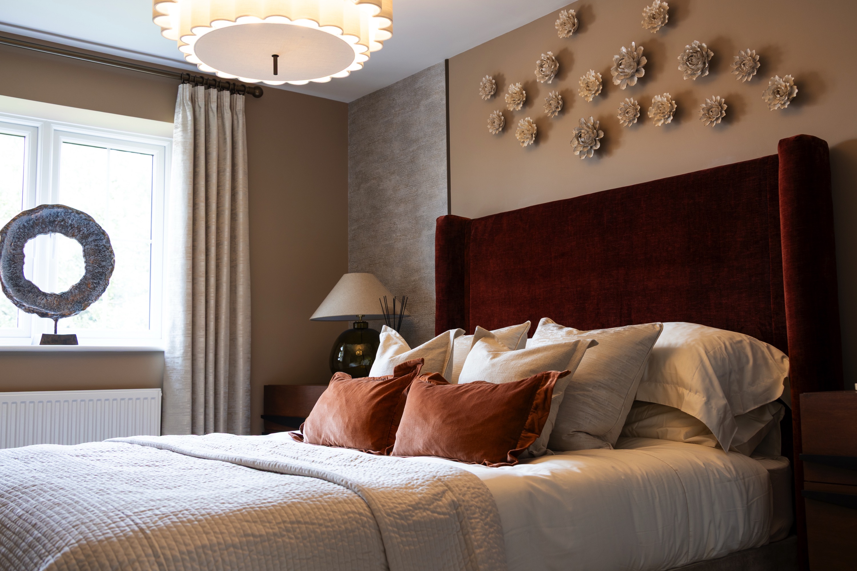 A cosy bedroom interior featuring a large bed with a plush velvet headboard in a deep red colour. The wall behind the bed is decorated with an array of 3D floral wall art. The bed is dressed with white bedding and styled with terracotta scatter cushions. A large window with curtains is visible on the left, with a unique ring-shaped decorative object on the windowsill.