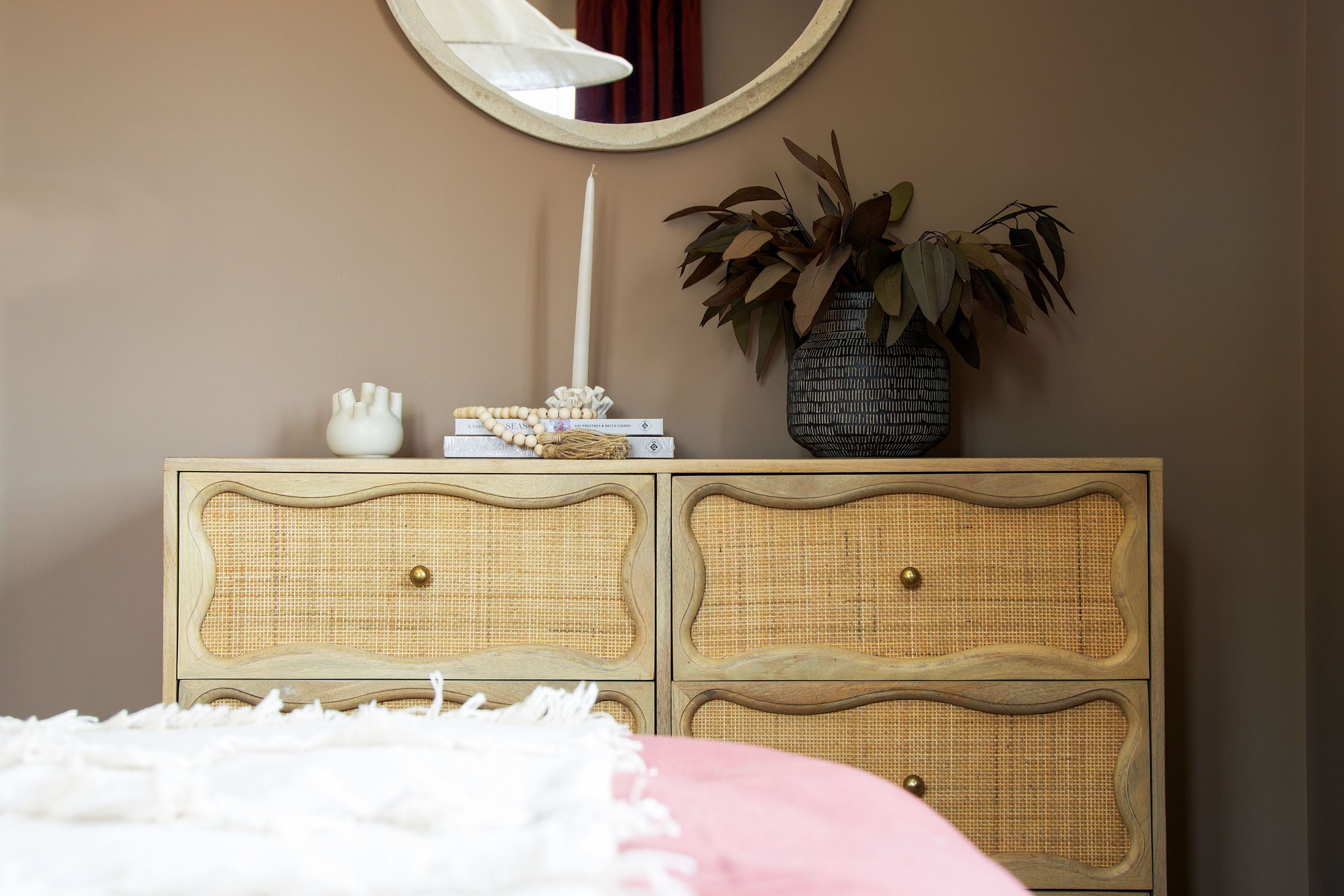 A detailed shot of a bedroom interior showcasing a chest of drawers with stylish rattan fronts. The top of the drawers is styled with various home decor items, including books, a vase of dried foliage, and a candle. A large round mirror hangs above on a warm-toned wall, with a section of the bed and its pink and white bedding visible in the foreground. The room features a natural colour palette.
