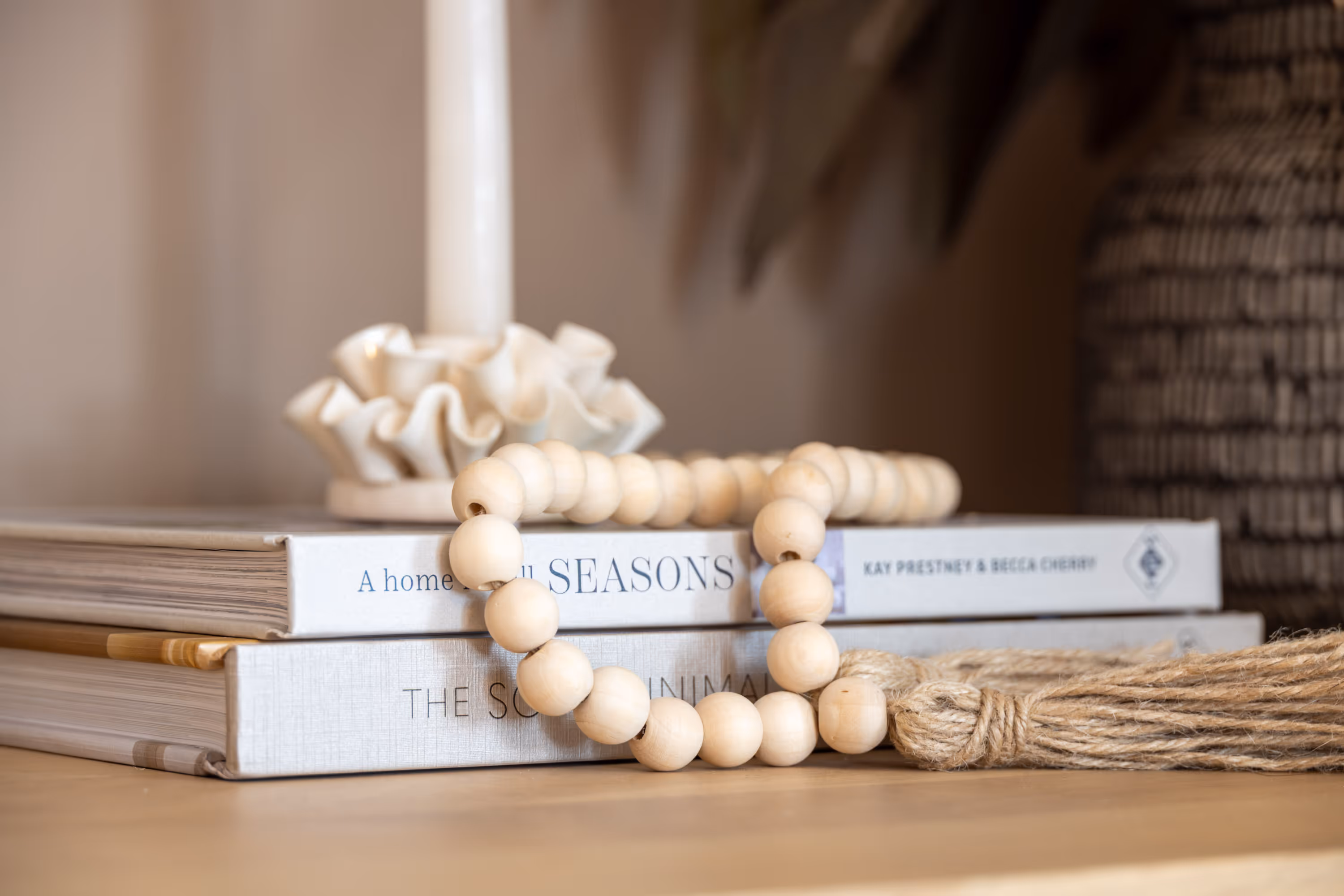 A close-up shot of a decorative arrangement on a wooden surface. In the foreground, a strand of light-colored wooden beads with a tassel rests on two stacked books with white covers. Behind the books, a white, ruffled ceramic candle holder with a tall white candle is visible. To the right, part of a woven basket and some blurred greenery complete the background.