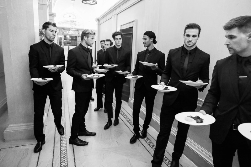 Yann Nury catering staff dressed in black suits holding plates with food standing in a hallway.