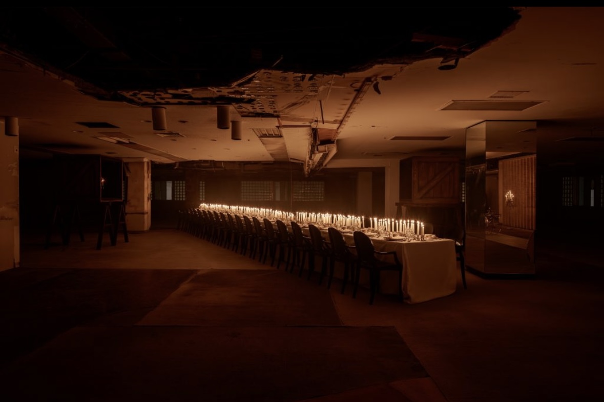 Catering event with long banquet table with white tablecloth set with plates and lit candles in a dimly lit industrial space.