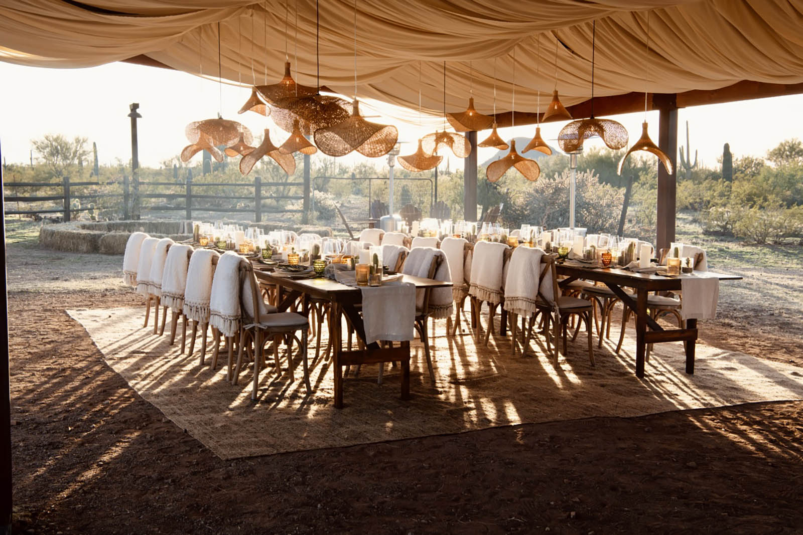 Private catering outdoor dining setup with a long wooden table and chairs covered with white blankets under a fabric canopy, surrounded by desert vegetation.