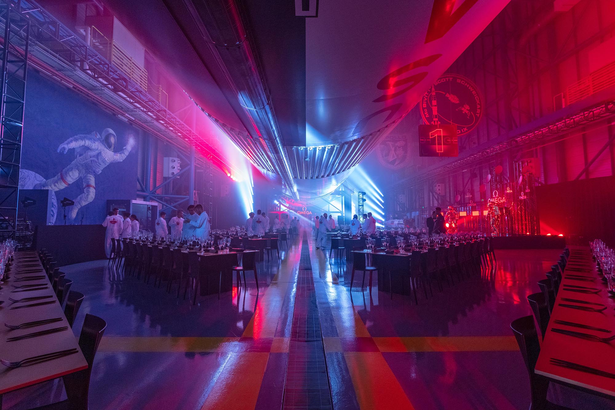 Nasa event space set for a dinner with rows of tables and chairs, colorful lighting, and a large rocket suspended overhead.