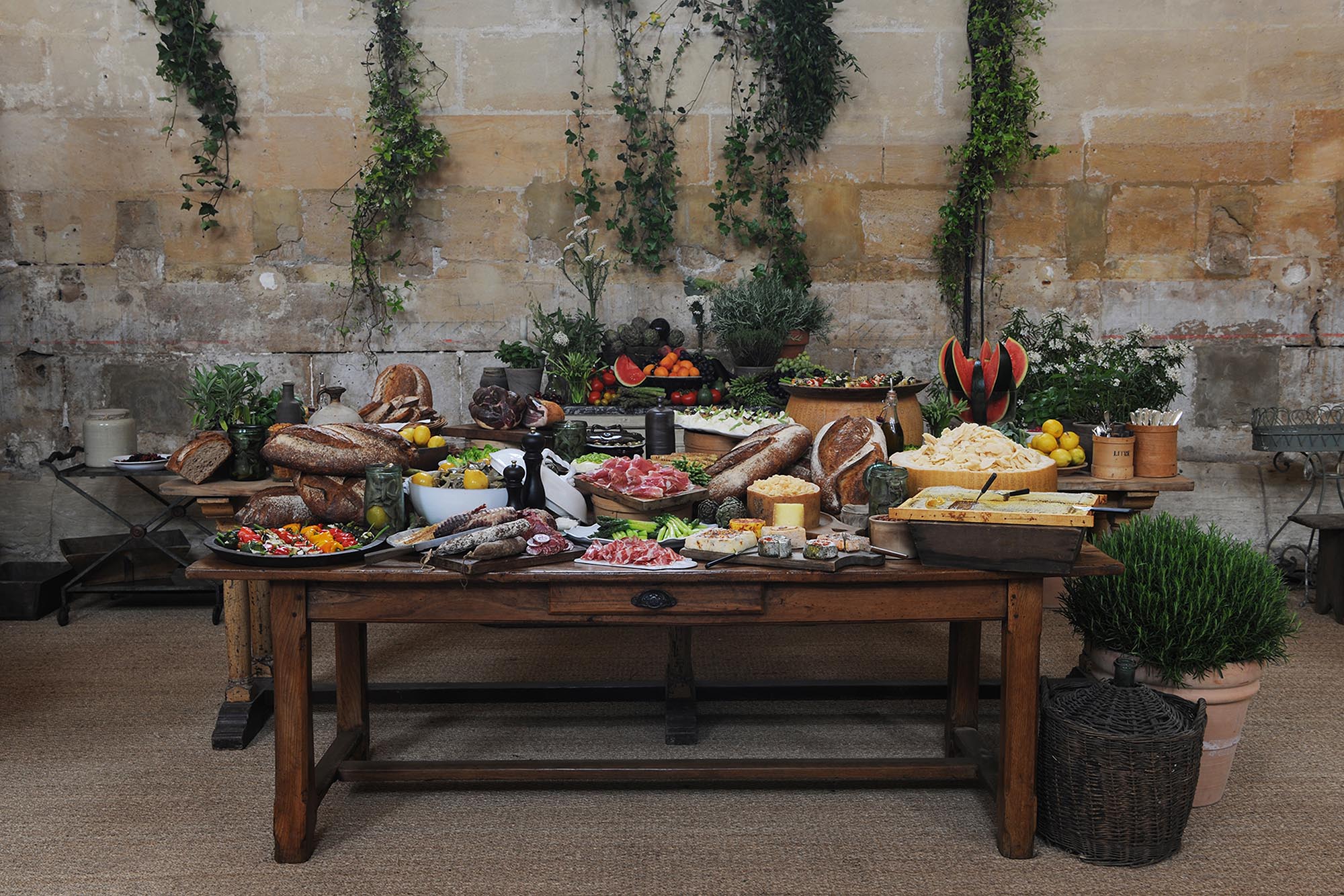 Dior Chantilly rustic buffet table with assorted breads, cured meats, cheeses, fresh vegetables, fruits, and herbs against a stone wall with hanging greenery.
