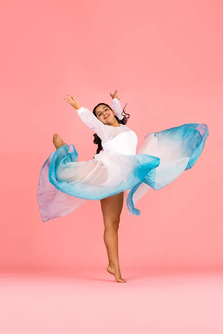 Smiling Danceology dancer in a flowing teal-and-white skirt on a coral backdrop