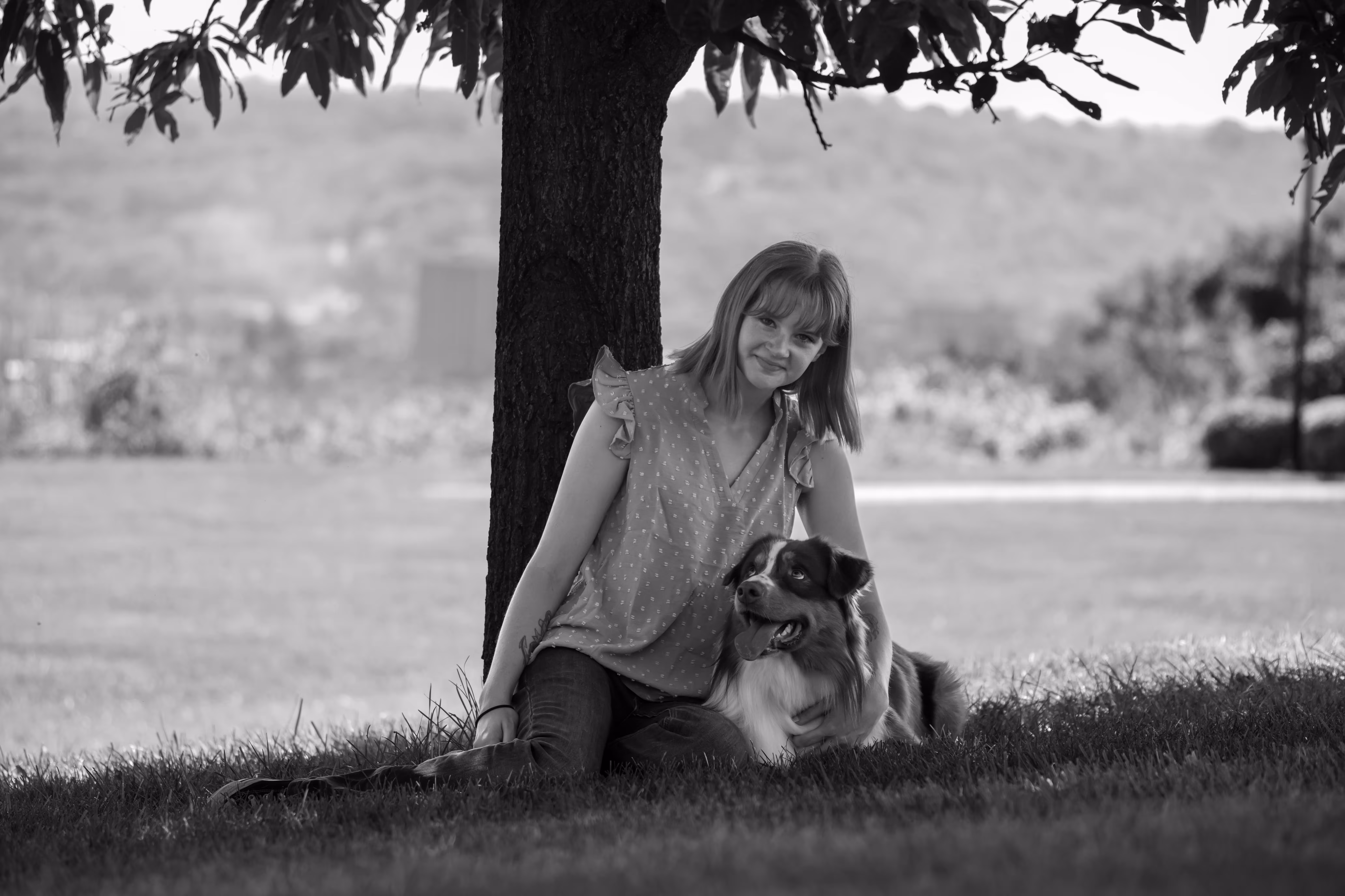 A black and white photograph of a young woman with shoulder-length hair sitting under a tree with her Australian Shepherd. She is wearing a sleeveless, ruffled blouse and jeans, smiling gently while resting her arm around her dog. The dog gazes off into the distance with a relaxed and content expression. The background features a soft-focus landscape of rolling hills and trees, adding depth to the serene outdoor scene.