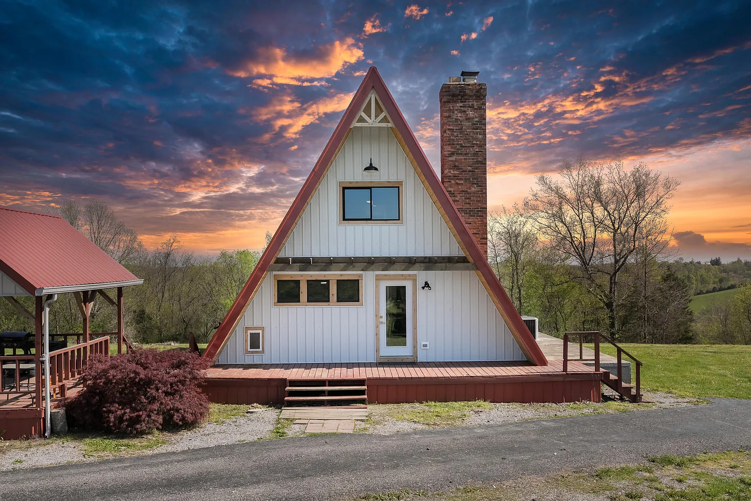 A-frame house with white siding, red trim, and a tall brick chimney at sunset with a colorful sky and surrounding trees.