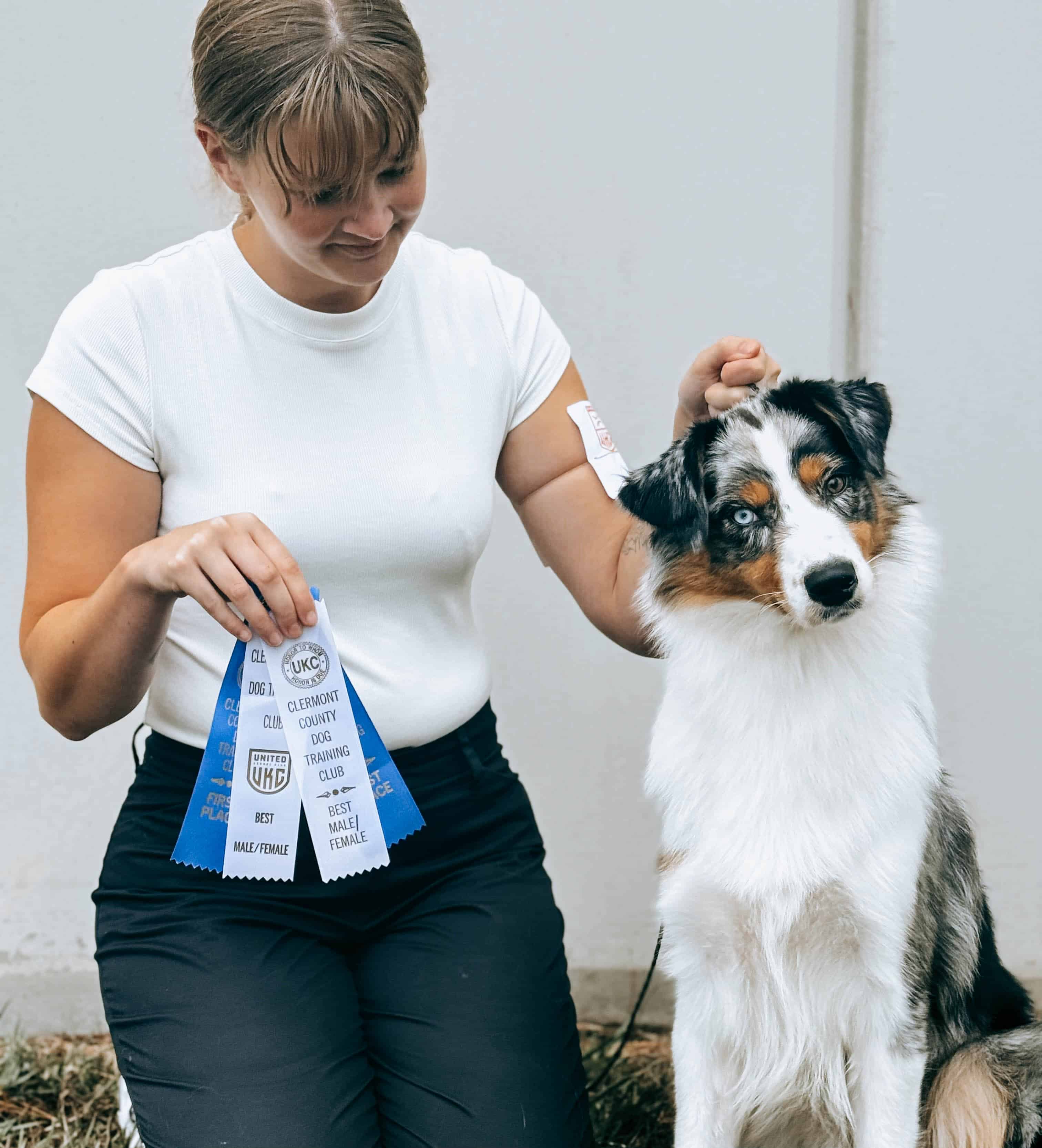 A woman kneels beside a blue merle Australian Shepherd holding multiple blue and white dog show ribbons, both looking proud after winning at a conformation event.