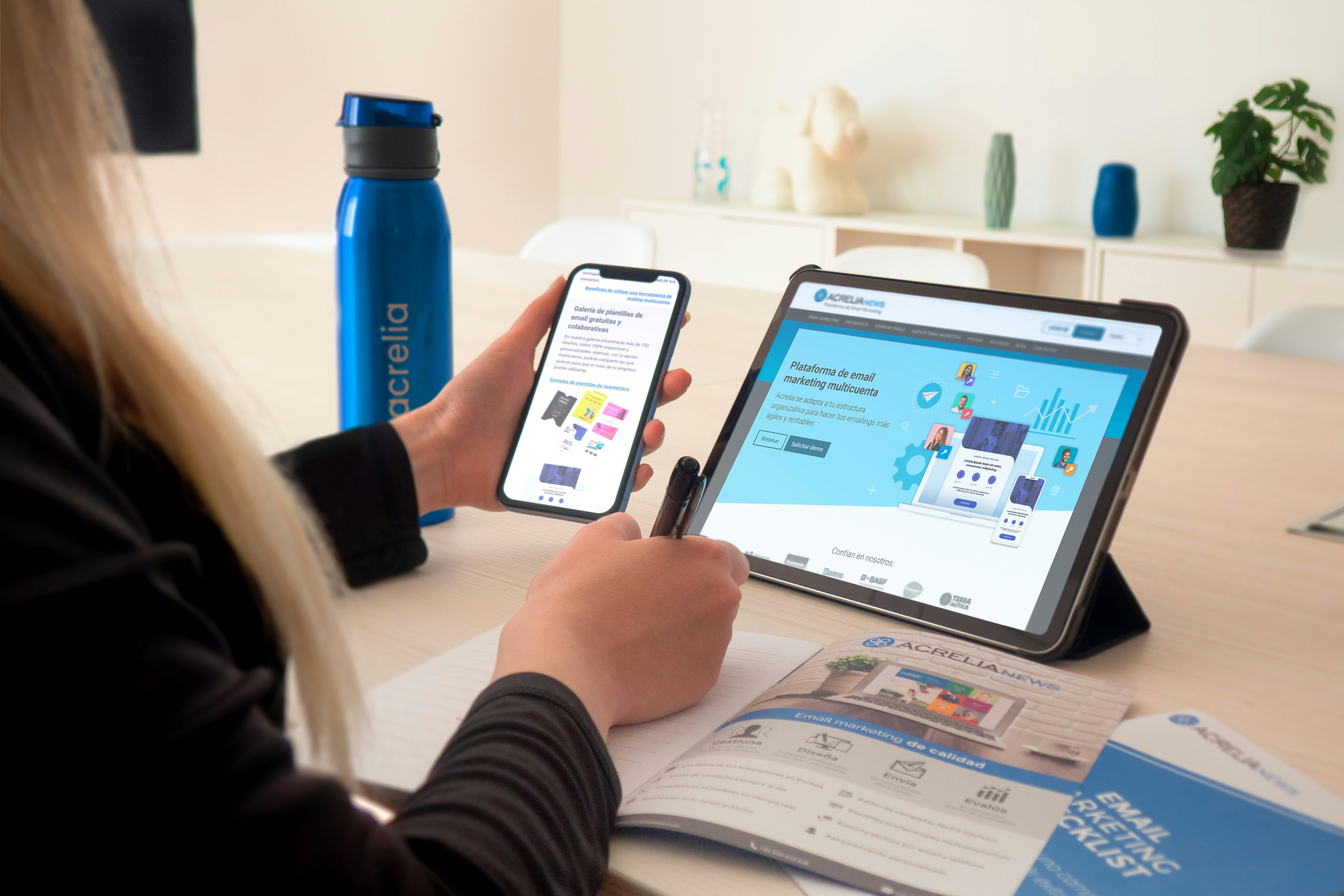 Person holding smartphone and writing notes with tablet displaying email marketing platform for campaign planning