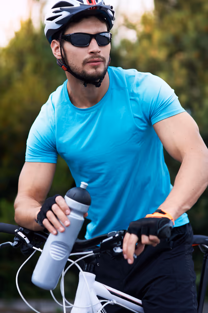 Cyclist in blue shirt, helmet, and sunglasses holding water bottle near bicycle