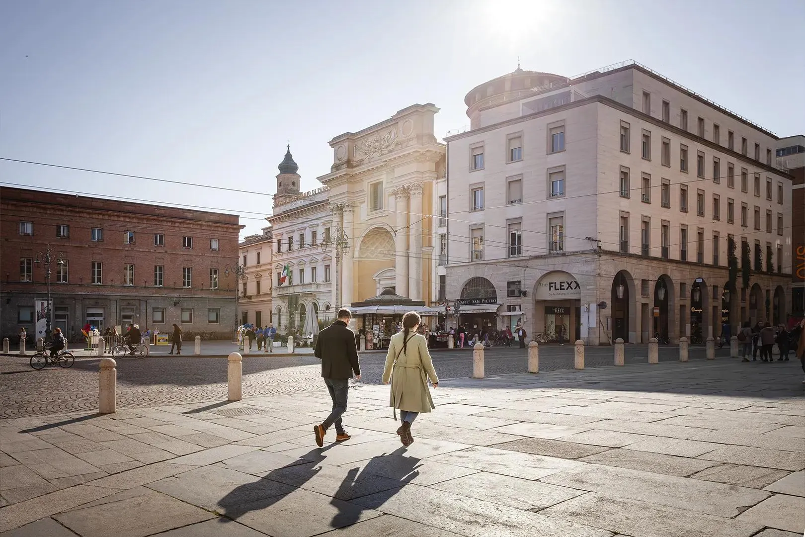 Due persone camminano in una piazza di Parma tra edifici storici e moderni.