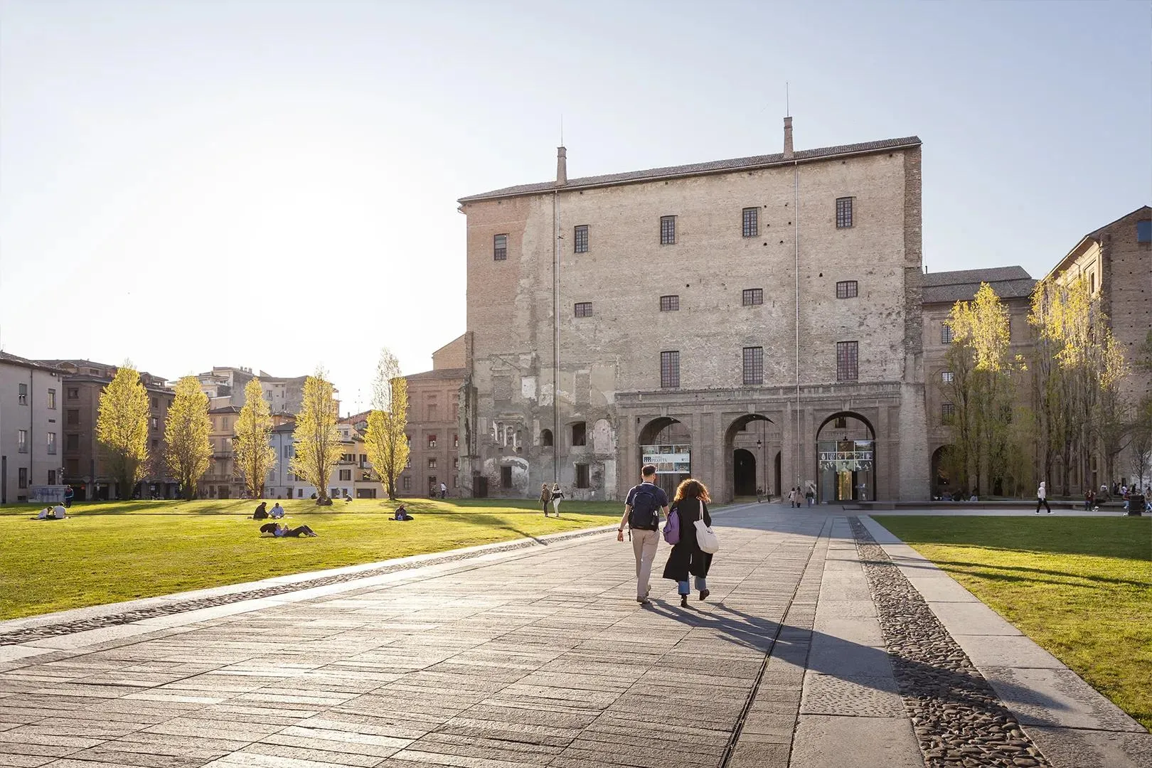 Piazza nel centro storico di Parma con edifici storici e passanti nel sole del pomeriggio.