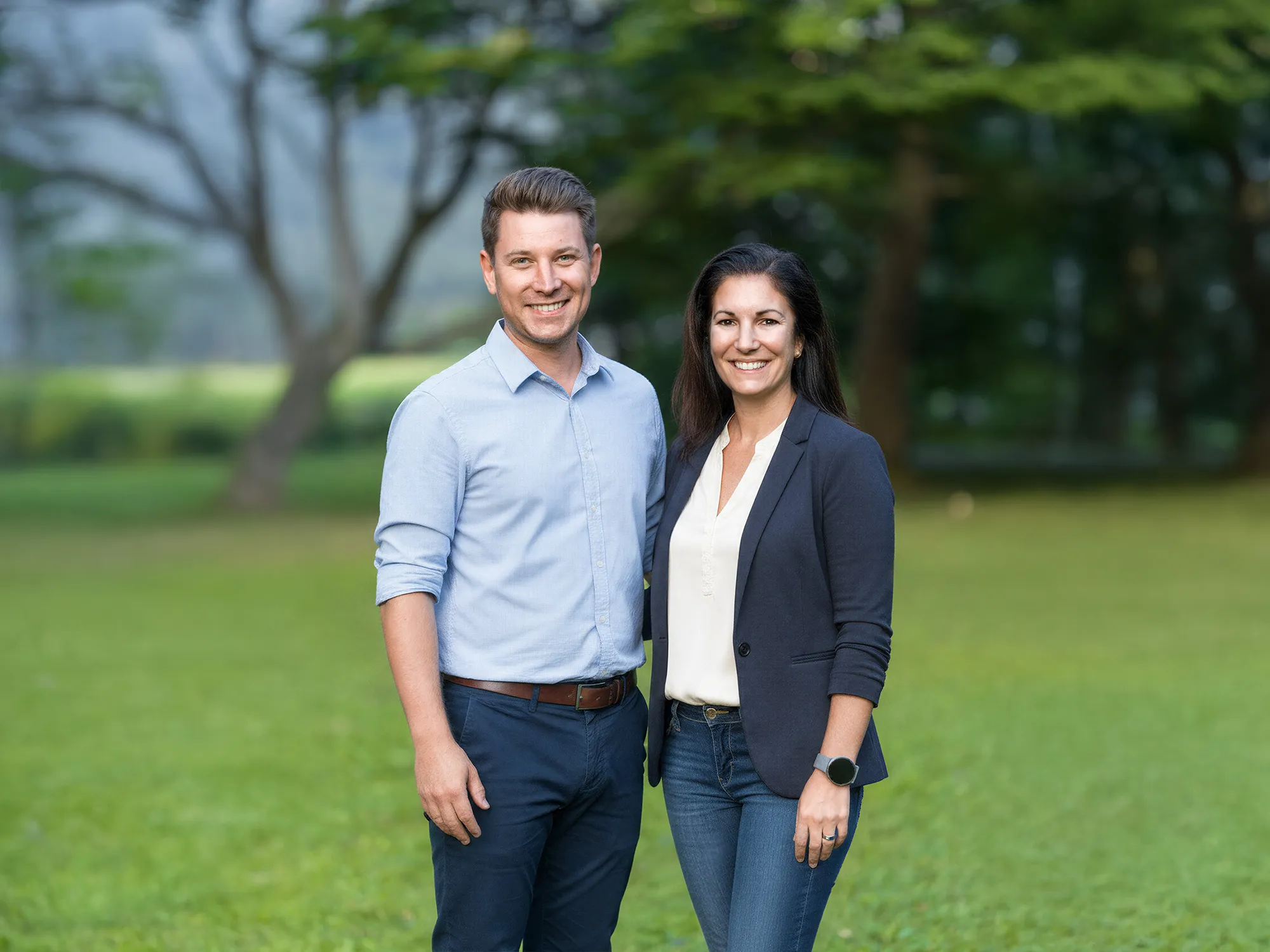 Die Gründer von Lehrlingsimpuls, Simon Schelkshorn und Heike Dormuth, stehen gemeinsam auf einer grünen Wiese. Beide lächeln in die Kamera, tragen Business-Casual Kleidung und wirken sympathisch und nahbar vor einem natürlichen Hintergrund mit Bäumen.