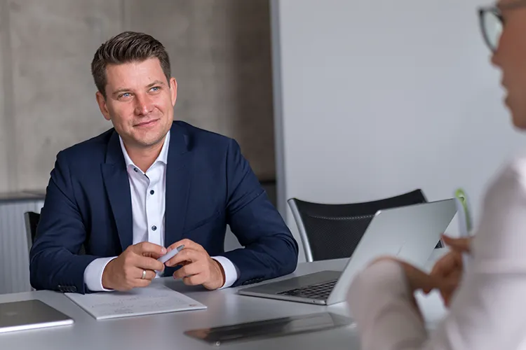 Simon Schelkshorn, Gründer von Lehrlingsimpuls, sitzt in einem modernen Besprechungsraum im Gespräch mit einer Person. Er trägt ein dunkelblaues Sakko und lächelt aufmerksam, während vor ihm Laptop und Unterlagen auf dem Tisch liegen.