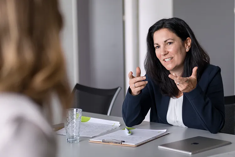 Heike Dormuth, Mitgründerin von Lehrlingsimpuls, sitzt in einem modernen Besprechungsraum und spricht engagiert mit einer Gesprächspartnerin. Vor ihr liegen Unterlagen, ein Glas Wasser und ein Laptop, während sie mit freundlicher Gestik ihre Gedanken erläutert.