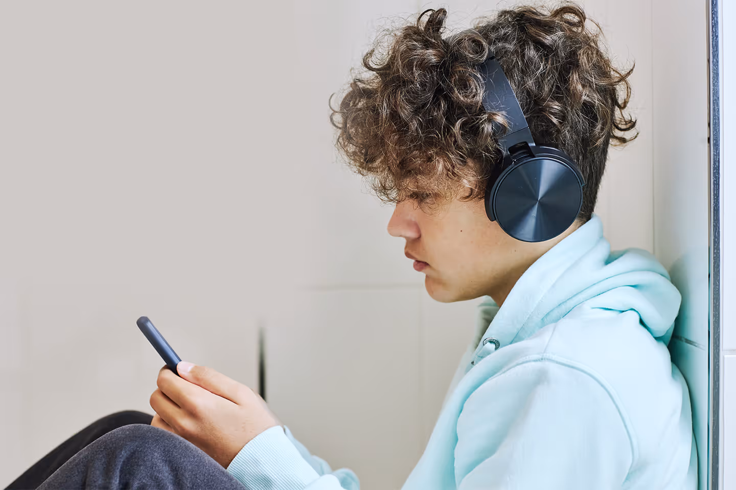 Young boy sat down leaning against a wall with headphones on holding a mobile phone.