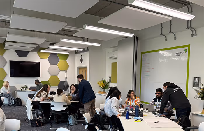 People in an office meeting room having discussions around tables with whiteboards and hexagonal wall decor.