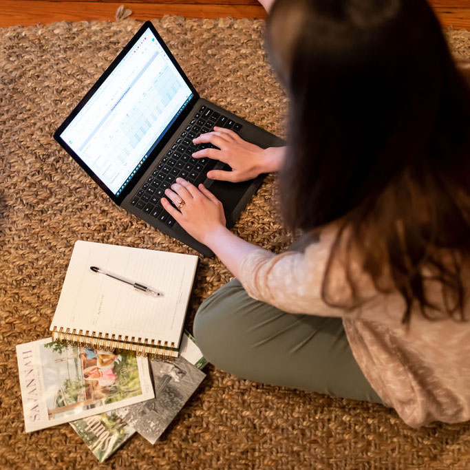 Woman sitting on a woven rug working on a laptop with spreadsheets open, surrounded by a notebook, pen, and printed design materials