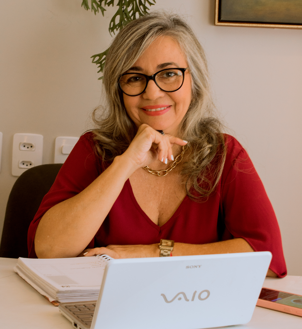 Paula Nicodemos sorrindo em um ambiente aconchegante. Usa blusa vermelha e trabalha em um notebook.