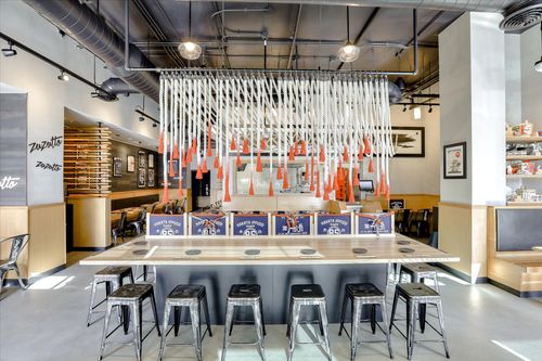 Modern Japanese restaurant interior with a long wooden table surrounded by metal stools and hanging white ropes with orange tassels above.
