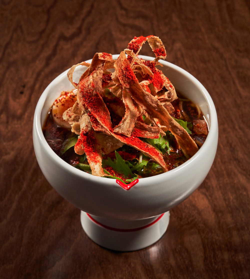 White bowl with a pedestal holding a spicy stew topped with crispy strips and green herbs on a wooden surface.