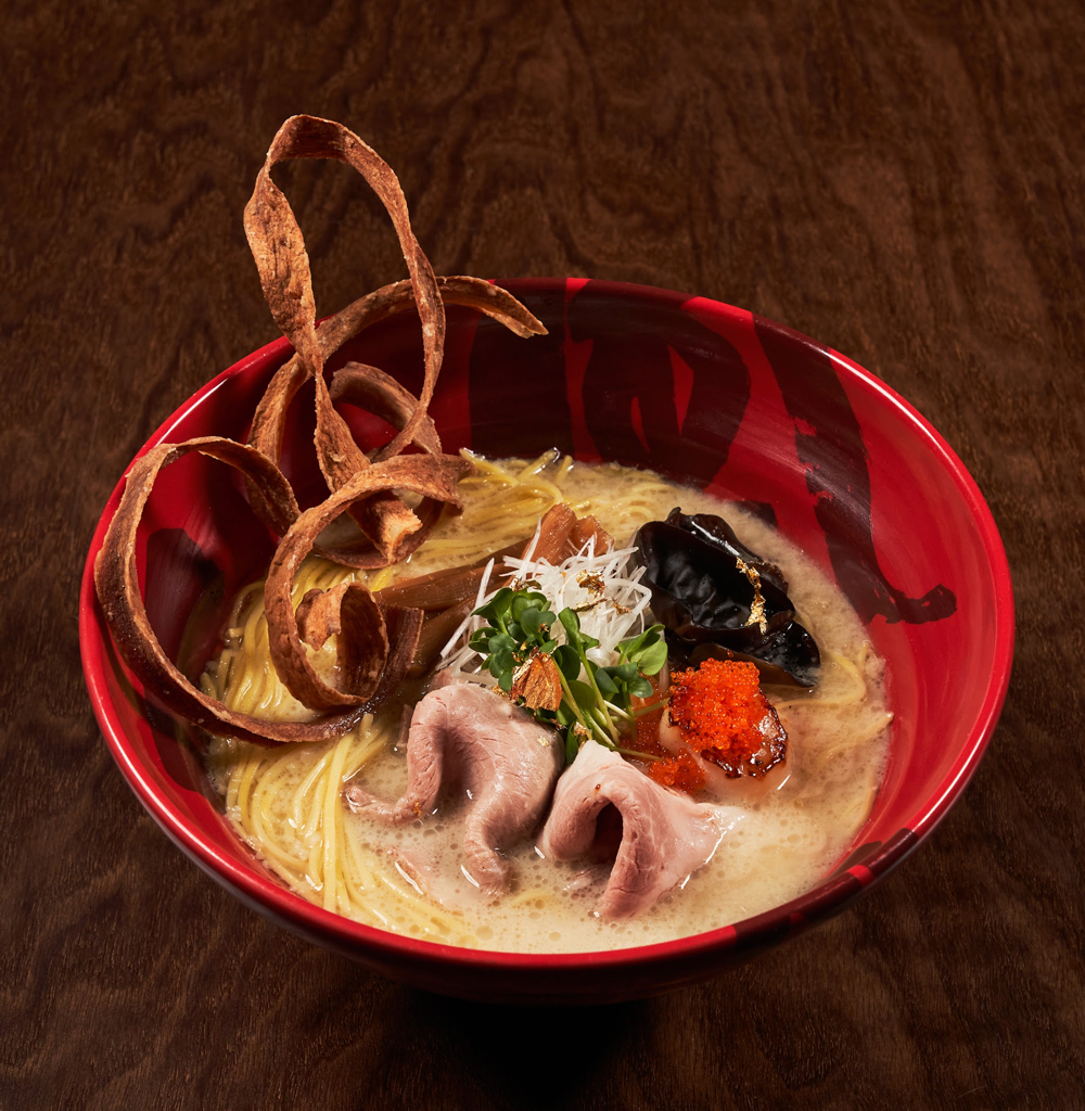Red bowl of tonkotsu ramen with creamy broth, noodles, slices of pork, black fungus, fish roe, greens, and crispy fried garnish on a wooden surface.