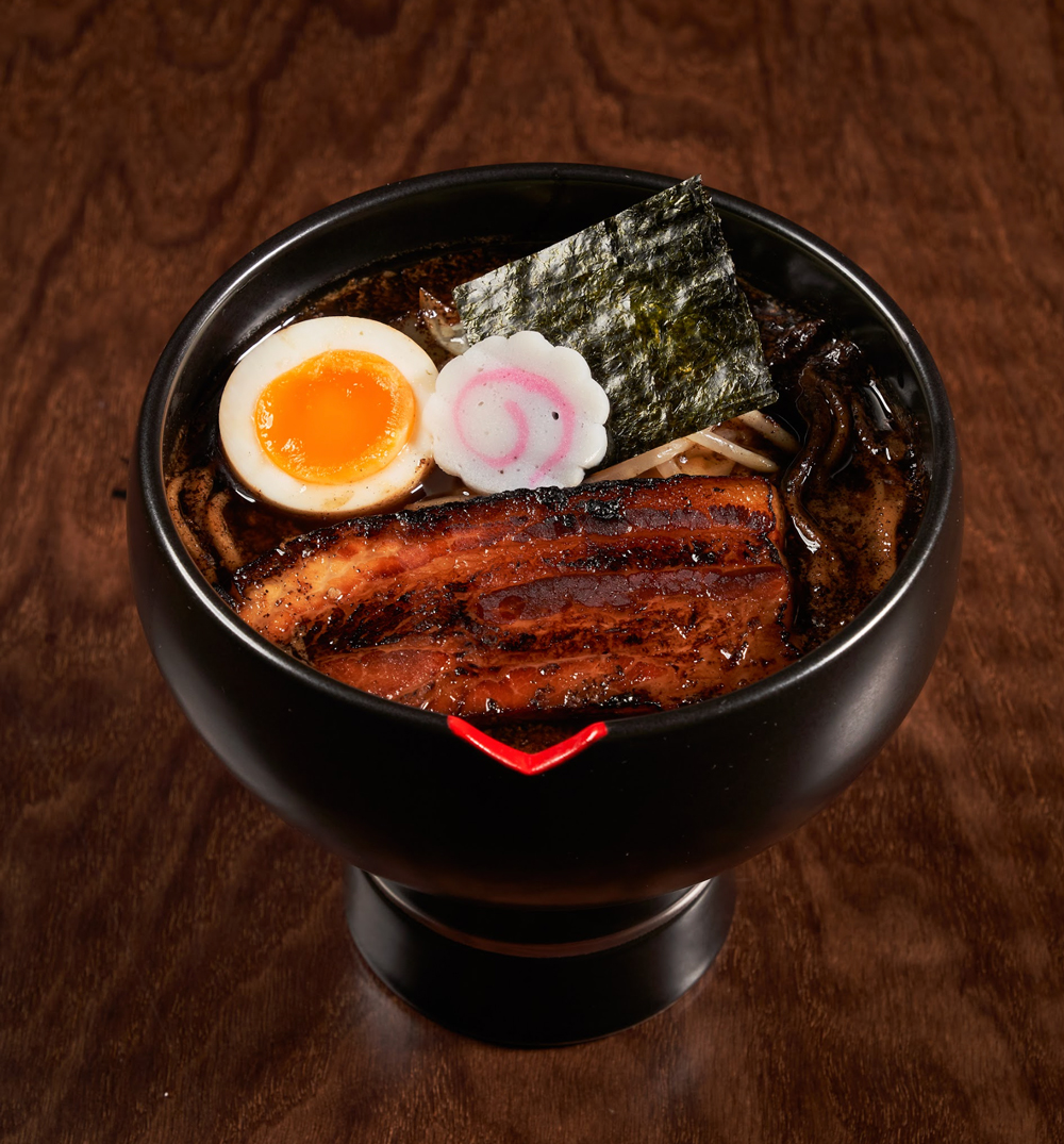 Black bowl of ramen with grilled pork belly, half soft-boiled egg, narutomaki, seaweed, and noodles in dark broth on a wooden surface.