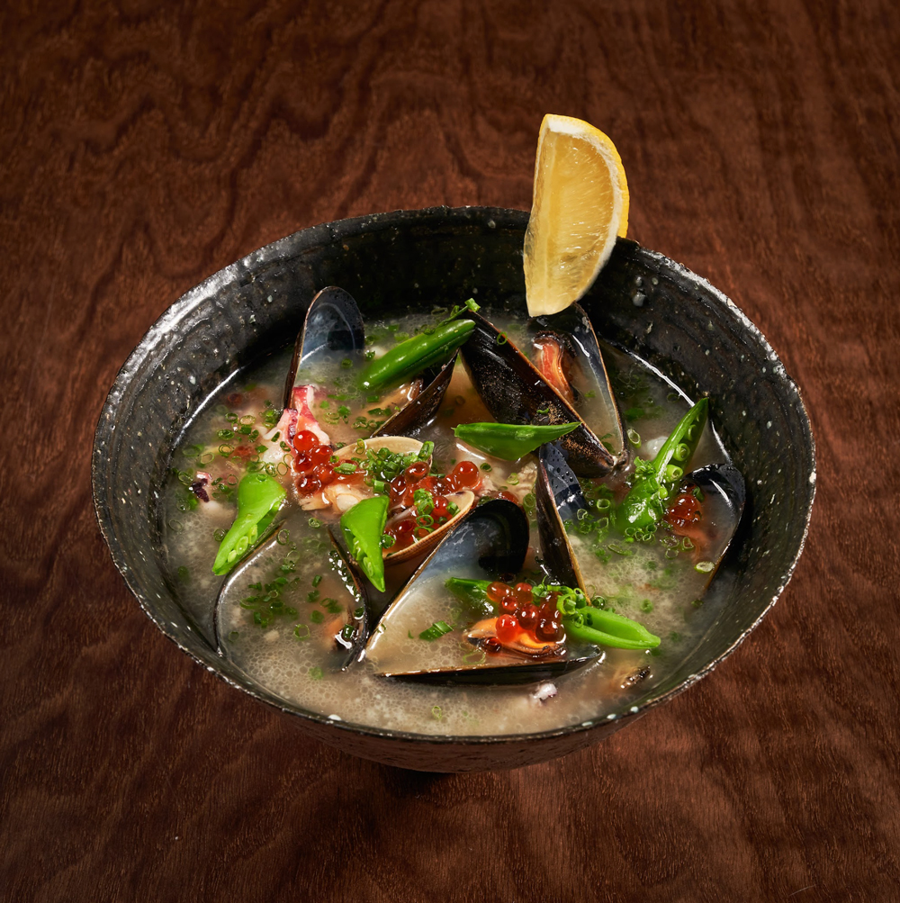 Black bowl of seafood soup with mussels, green peas, red roe, chopped green onions, and a lemon wedge on the rim, set on a wooden surface.