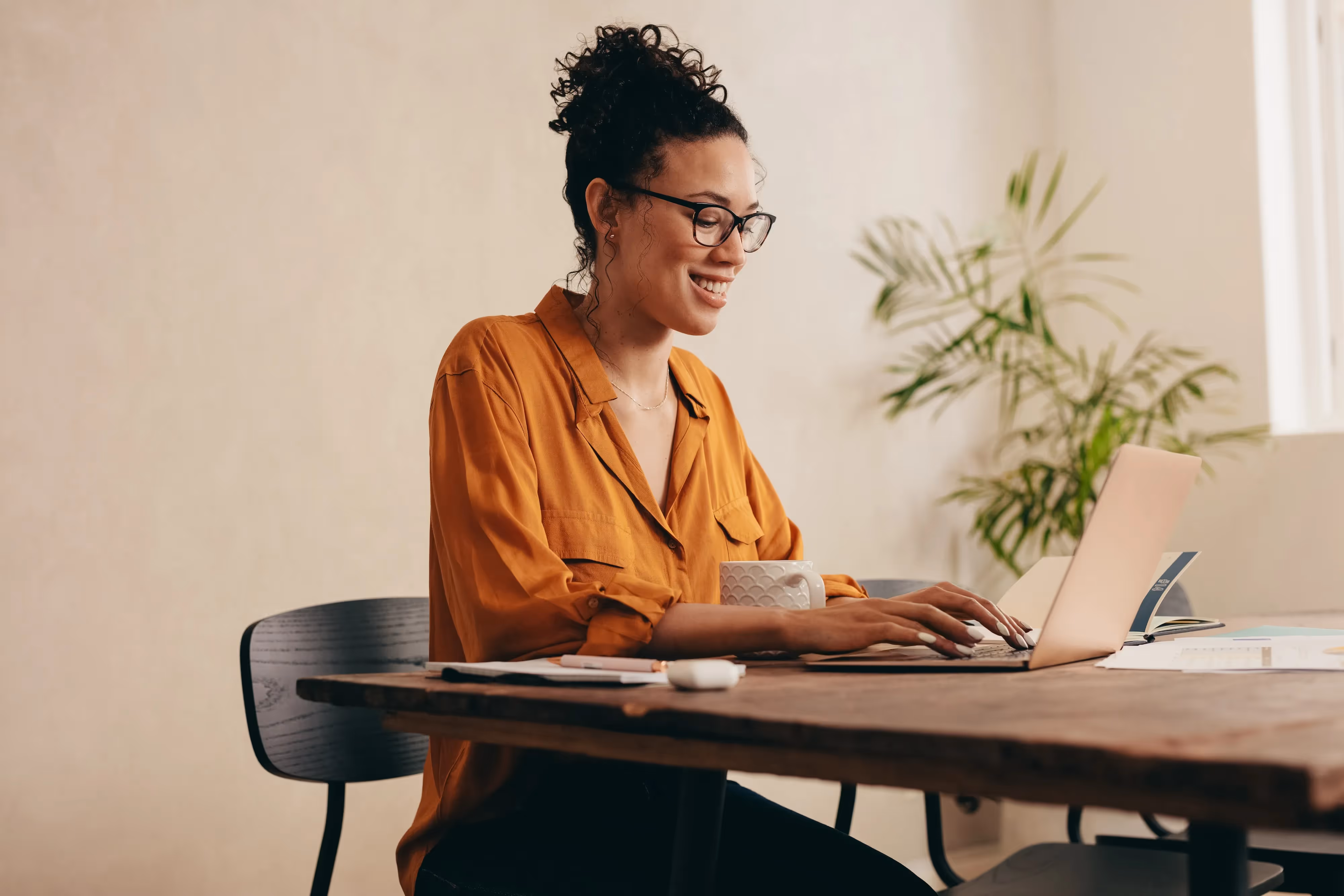 Smiling woman wearing glasses working on a laptop at a wooden table, depicting a comfortable work-from-home or co-working environment near Crossing at 9th in Salt Lake City.