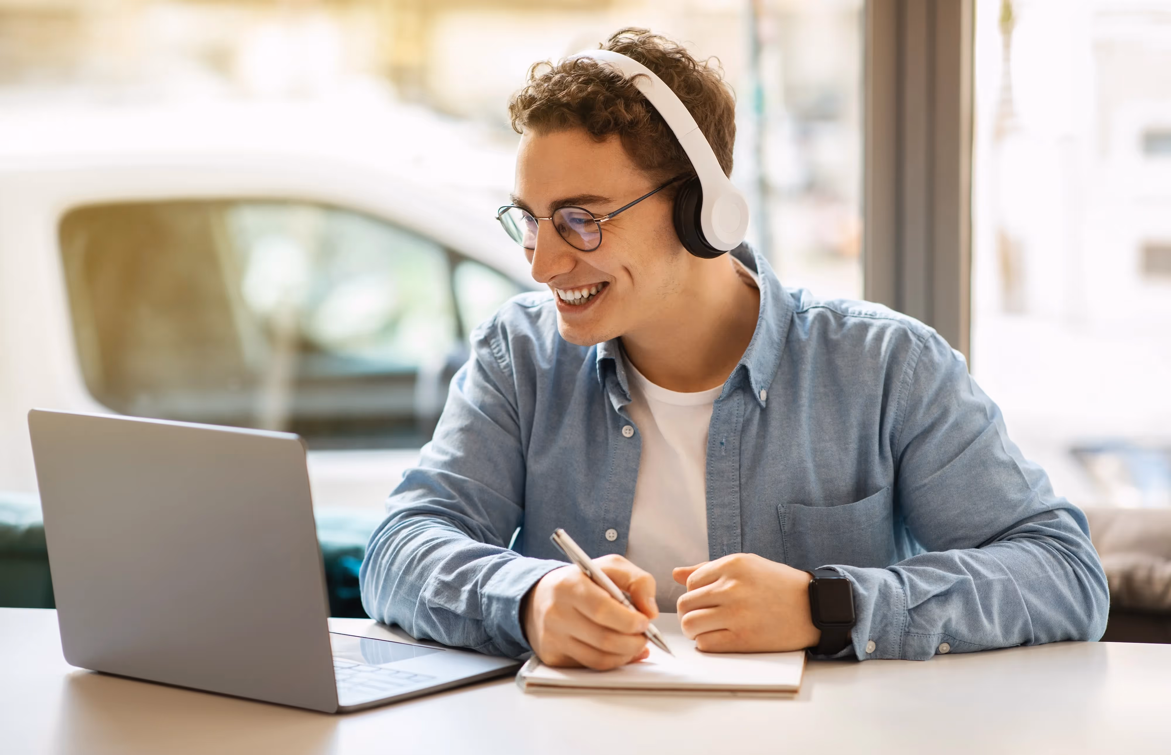 Smiling male student wearing headphones and glasses, writing in a notebook while using a laptop, representing a productive study environment near Crossing at 9th in Salt Lake City, Utah.