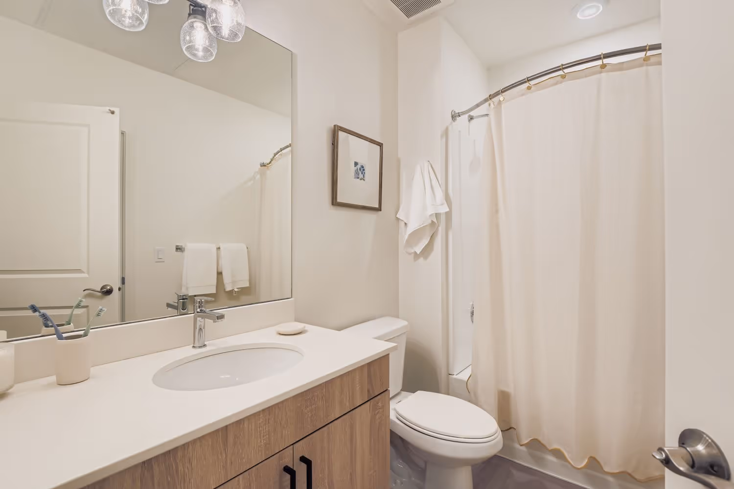 Modern bathroom with a single vanity, wood-grain cabinets, toilet, and a bathtub with a cream-colored shower curtain at Crossing at 9th in Salt Lake City, Utah.