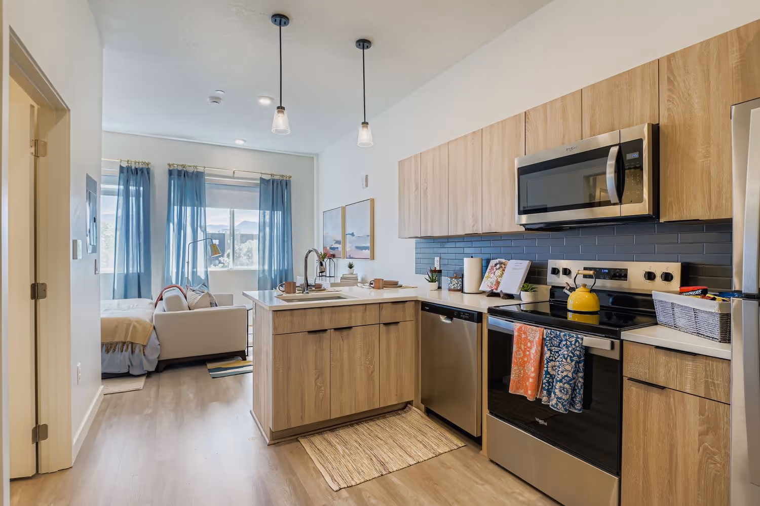 Apartment kitchen featuring light wood-grain cabinets, subway tile backsplash, and stainless steel appliances, with a view into the living area, at Crossing at 9th in Salt Lake City.