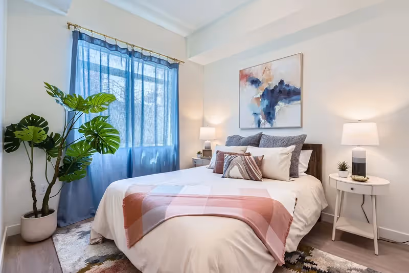 Bright bedroom with a large bed, blue curtains, and modern art, showcasing an apartment interior at Crossing at 9th in Salt Lake City.