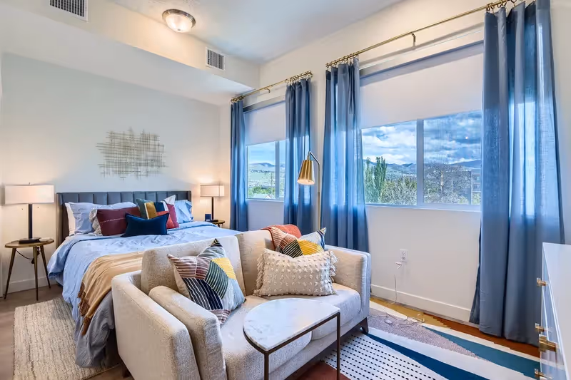 Spacious bedroom with a bed, small sofa, and large windows offering scenic views, in an apartment at Crossing at 9th in Salt Lake City.