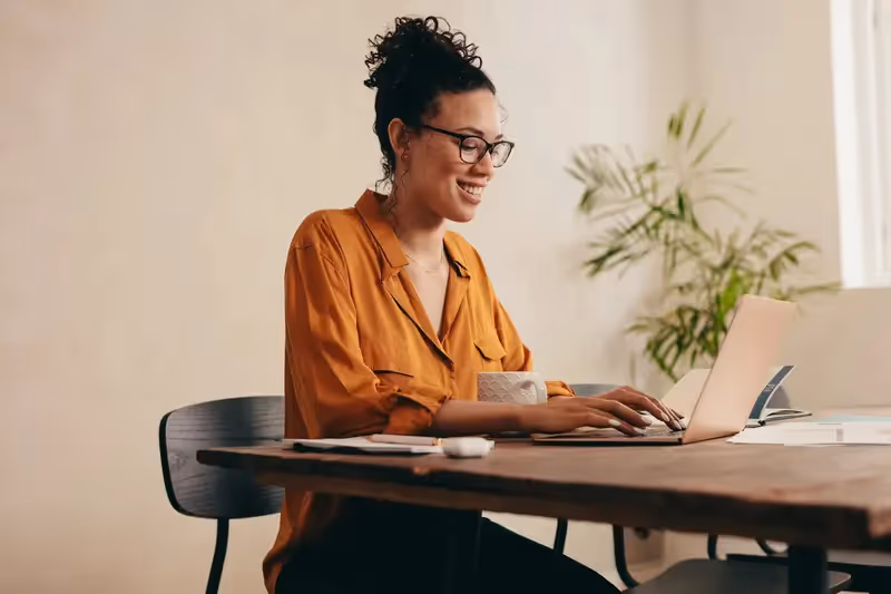 Smiling woman wearing glasses working on a laptop at a wooden table, depicting a comfortable work-from-home or co-working environment near Crossing at 9th in Salt Lake City.
