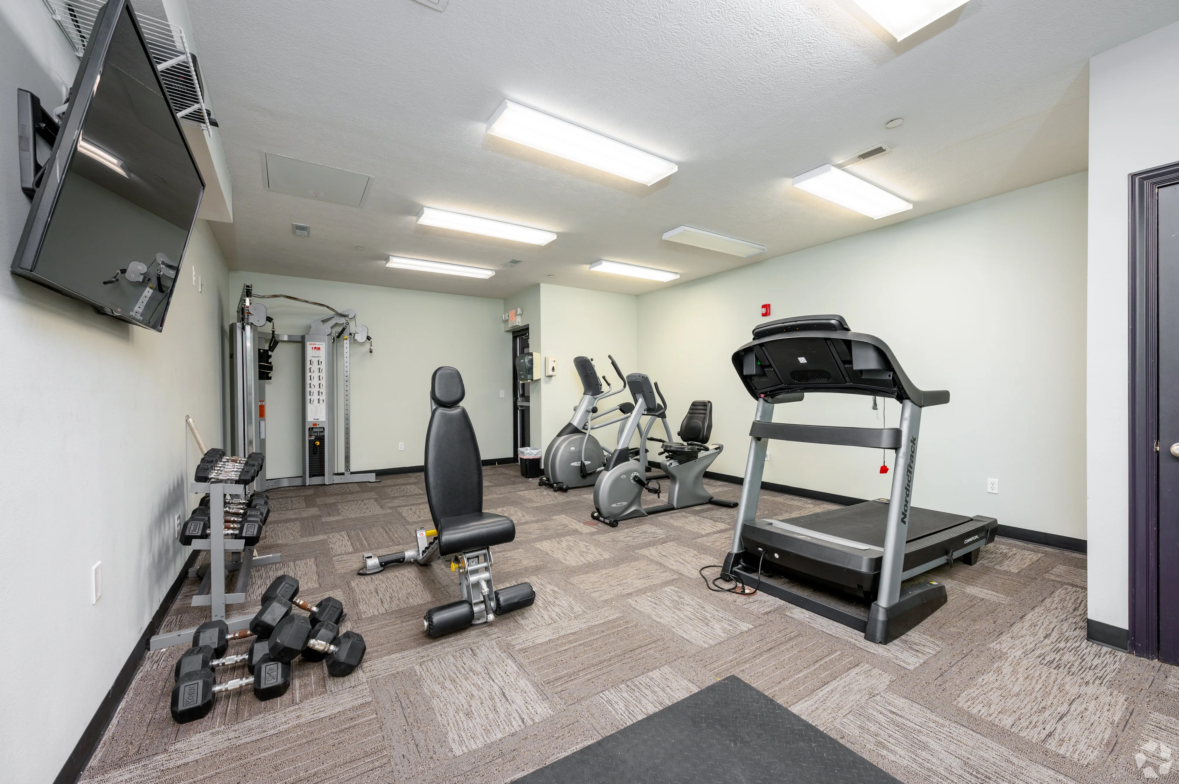 On-site fitness center at 7 Ten featuring treadmills, ellipticals, weight bench, and dumbbells with a wall-mounted TV.