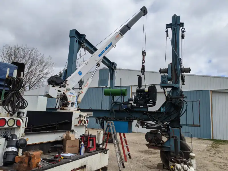 Heavy-duty field service truck with a crane hoisting a large engine assembly from an industrial machine during an outdoor equipment repair.