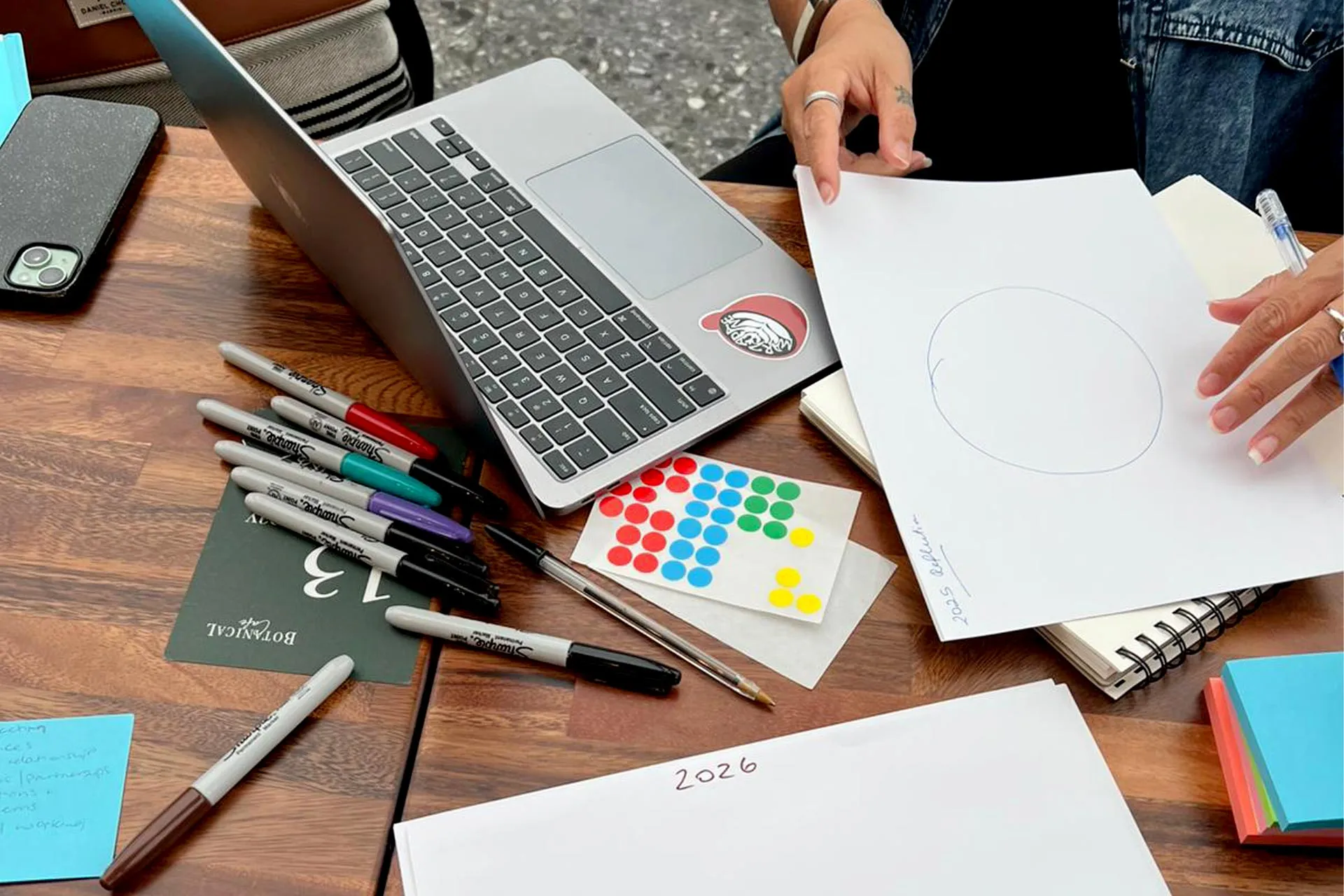 Person holding a sheet of paper with a circle drawing next to a laptop, surrounded by colored Sharpie markers and sticky notes on a wooden table.