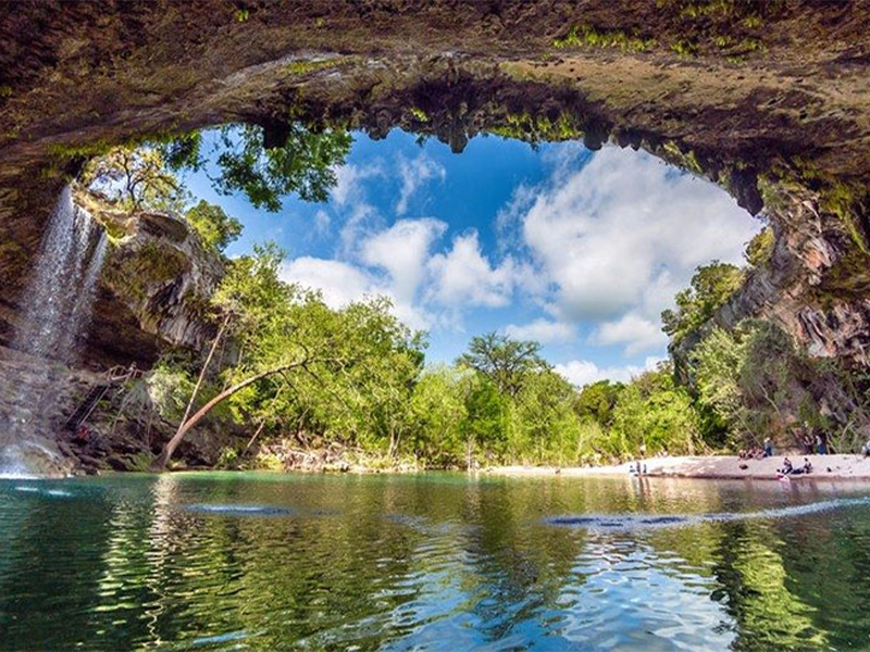 A serene natural scene in Dripping Springs Texas featuring cascading water droplets from a rocky overhang into a calm, clear blue pool surrounded by greenery.
