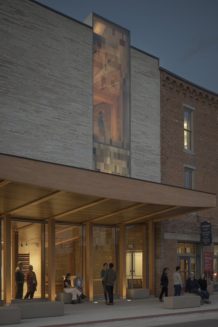 The exterior close up of the building with people gathering outside on benches and a large full-length window on the 2nd floor that a person is looking out of.