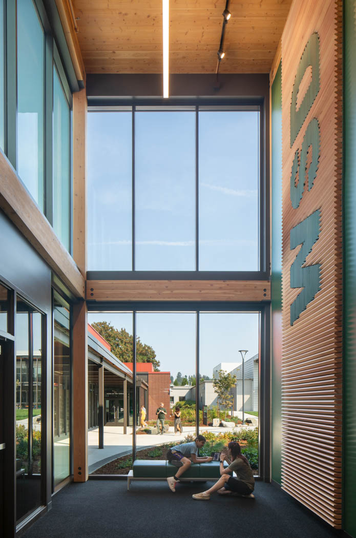 A vertical image of inside the building in a corner showing the side of the building from a window view and people are hanging out on the benches and walking outside. Exposed mass timber is on the side walls. And a symmetrical image of the beams and columns. 