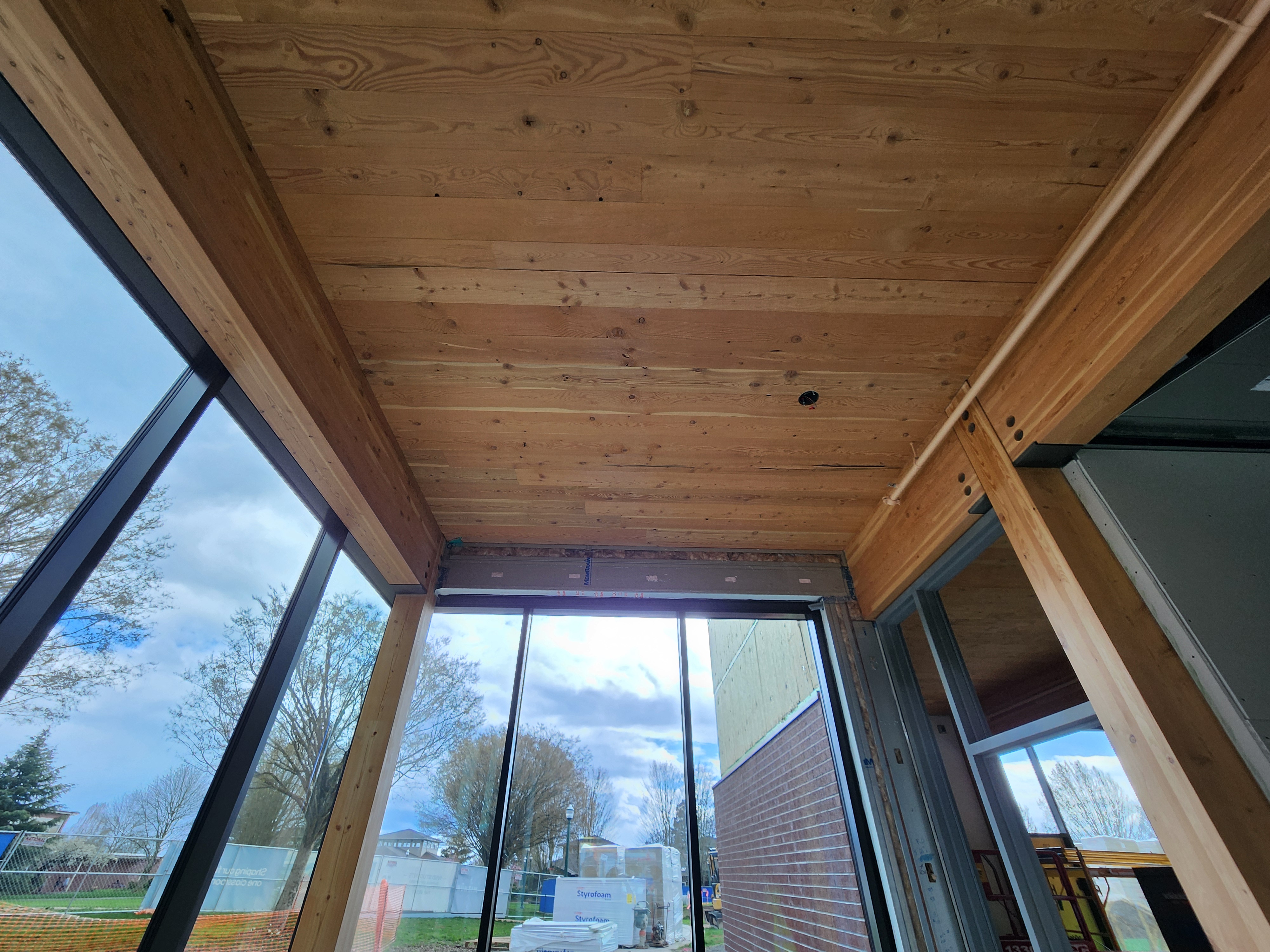 A corner space at the end of the building with floor to ceiling windows of a spring looking day. Wooden mass timber beams and columns and a mass timber ceiling exposed inside. 